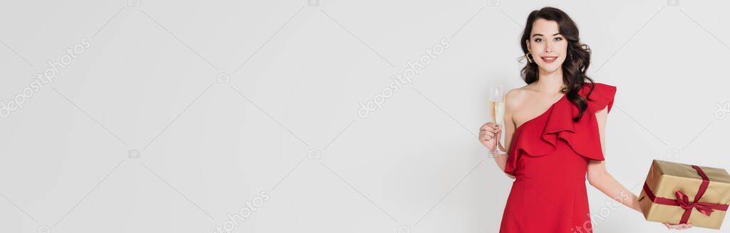 Brunette woman in red dress smiling while holding present and glass of champagne isolated on grey, banner 