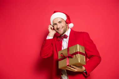 Noel Baba şapkalı gülümseyen adam kulaklıkla müzik dinliyor ve kırmızı arka planda hediyesini tutuyor. 