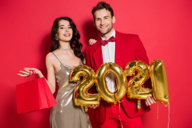 Elinde 2021 numara şeklinde balonlar tutan gülümseyen adam kırmızı arka planda alışveriş torbaları olan kız arkadaşının yanında.