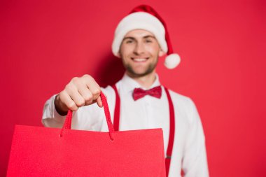 Kırmızı arka planda bulanık arka planda, Noel Baba şapkalı gülümseyen bir adamın elindeki alışveriş torbaları.