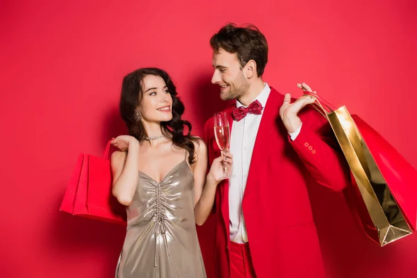 Casal Elegante Com Copo Champanhe Sacos Compras Sorrindo Fundo Vermelho — Fotografia de Stock