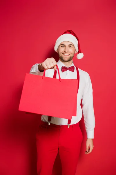 Stylischer Mann Mit Weihnachtsmütze Lächelt Während Einkaufstüten Auf Rotem Hintergrund — Stockfoto
