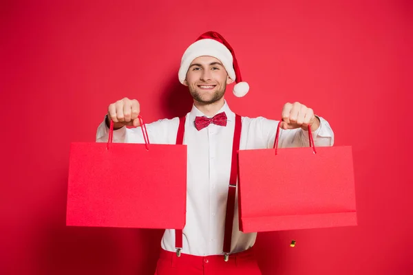 Junger Mann Mit Weihnachtsmannmütze Hält Einkaufstüten Der Hand Und Lächelt — Stockfoto