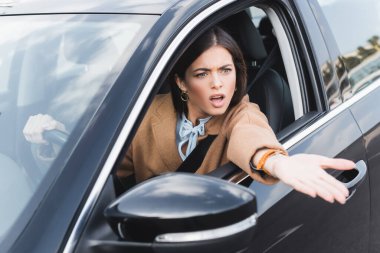 irritated woman shouting and pointing with hand from side window while driving car on blurred foreground clipart