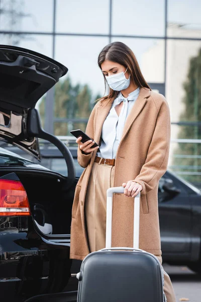 트렌치코트를 의료용 마스크를 여성이 트렁크 근처에서 가방을 스마트폰으로 메시지를 보내고 — 스톡 사진