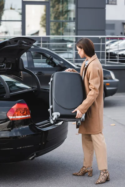 young woman in stylish autumn clothes loading suitcase in car trunk