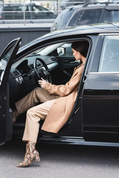 ドアを開けてハンドルを握って車の中に座っているスタイリッシュな服装の若い女性 — ストック写真