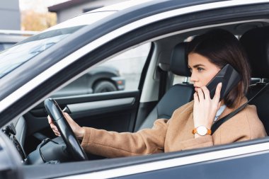 Genç bir kadın araba kullanıyor ve cep telefonuyla konuşuyor.