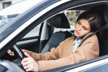 Sonbahar kıyafetli şık bir kadın araba kullanıyor ve cep telefonuyla konuşuyor.