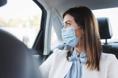 Tıbbi maskeli şık bir kadın, bulanık arka planda arabanın arka koltuğunda otururken camdan dışarı bakıyor.