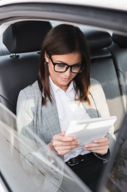 Gözlüklü mutlu iş kadını, bulanık ön planda arabada giderken dijital tablet kullanıyor.