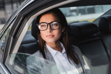 young businesswoman in eyeglasses looking out from open window while riding in car on blurred foreground clipart