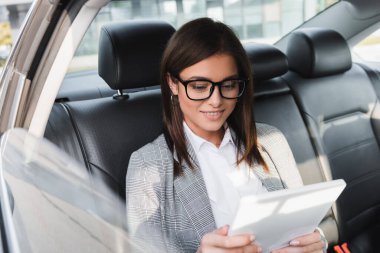 Aracın arka koltuğunda bulanık ön planda giderken dijital tablet kullanan pozitif iş kadını