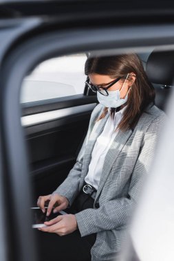 Tıbbi maskeli bir iş kadını, bulanık ön planda arabada giderken dijital tablet kullanıyor.