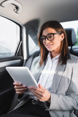 positive businesswoman using digital tablet while riding in car on back seat clipart