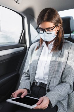 Arabada dijital tablet kullanan gözlüklü ve tıbbi maskeli bir iş kadını.