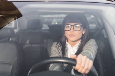 Resmi giyimli ve gözlüklü genç bir iş kadını bulanık ön planda araba kullanıyor.