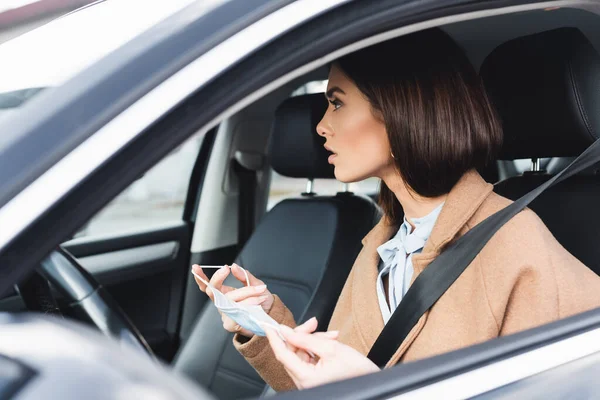 Jovem Mulher Olhando Para Longe Enquanto Sentado Carro Segurando Máscara — Fotografia de Stock