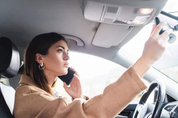 車の中でスマホで会話しながらバックミラーを調整する若い女性 — ストック写真