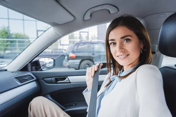 車に座ってシートベルトを固定しながらカメラで微笑むエレガントな女性 — ストック写真