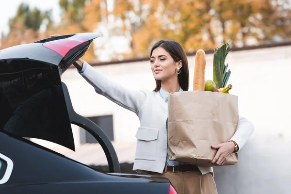 Uśmiechnięta Kobieta Otwiera Bagażnik Samochodu Trzymając Torbę Zakupy Jedzeniem — Zdjęcie stockowe