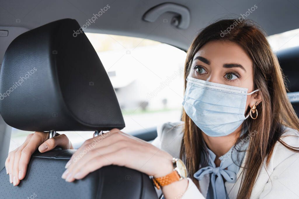 young woman in medical mask touching front seat and looking ahead while riding in car