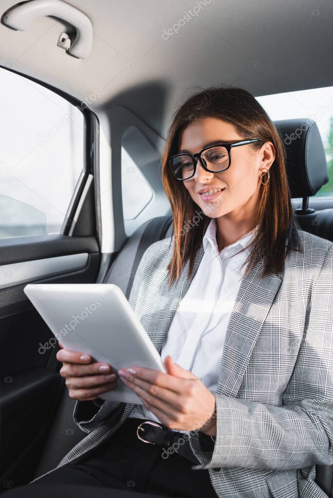 positive businesswoman using digital tablet while riding in car on back seat