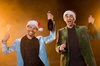 Noel Baba şapkalı heyecanlı adamlar ellerinde şampanya şişeleri ve siyah bardaklarla 