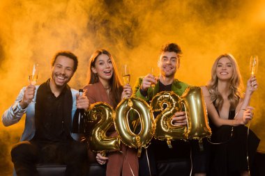 Mutlu ırklar arası arkadaşlar ellerinde şampanya bardakları ve 2021 numaralı balonlarla turuncu renkli dumanlı.