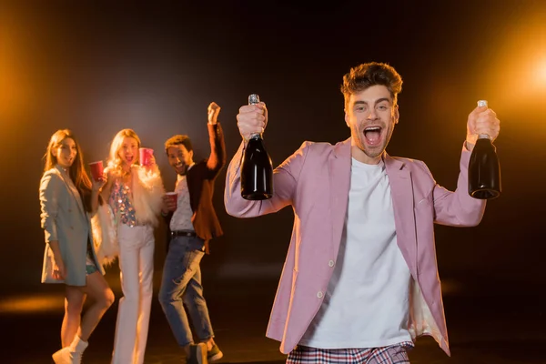 Excited Man Holding Bottles Champagne Multicultural Friends Blurred Black Background — Stock Photo, Image