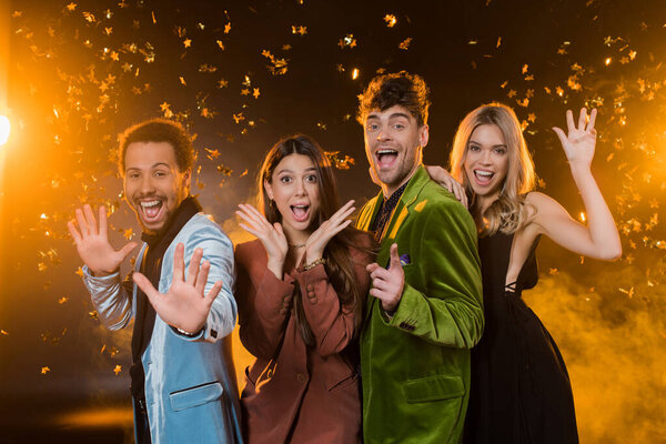 excited interracial friends gesturing near falling confetti on black