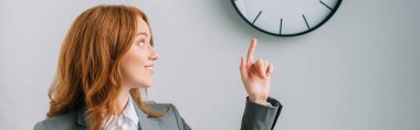 Positive redhead businesswoman pointing with finger and looking at wall clock on grey, banner clipart