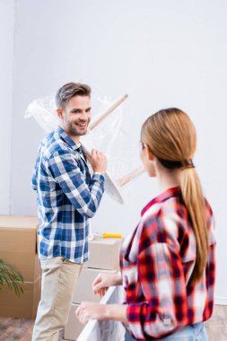 Kahve masası polietilen kaplı mutlu genç adam, evde bulanık ön planda bir kadına bakıyor.