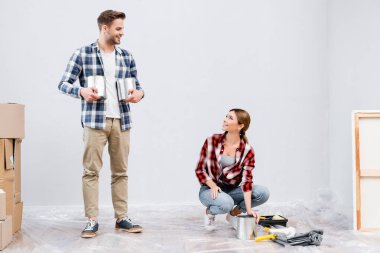 Boyalı teneke kutularla gülümseyen genç bir adam. Evdeki boya rulolarının yanında oturan kadına bakıyor.