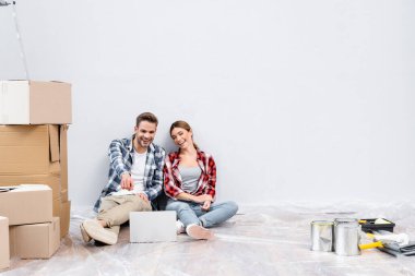 Evdeki karton kutuların yanında dizüstü bilgisayara bakıp gülen mutlu genç bir çift.
