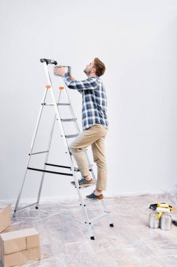 full length of young man with pot standing on ladder under leaking ceiling at home clipart