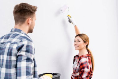 happy young woman with roller painting wall while looking at man indoors clipart