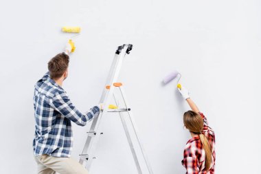 back view of young couple with rollers painting wall near ladder isolated on white  clipart