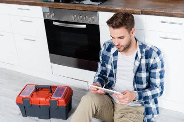 Genç adam, mutfak zemininde alet çantası ve fırının yanında otururken tablete bakıyor.