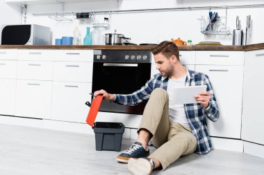Tabletli genç adam evde yerde otururken boş alet çantasına bakıyor.