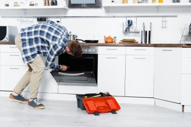 full length of young man with pliers bending down near oven and toolbox in kitchen clipart