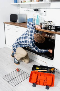 full length of young man repairing oven while sitting on floor near grid and toolbox in kitchen clipart