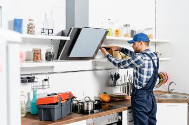 young handyman using drill near extractor fan on blurred foreground in kitchen clipart