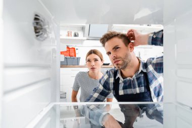 thoughtful handyman and young woman looking at freezer on blurred foreground in kitchen clipart