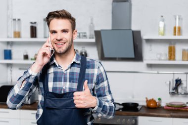 Arka planda bulanık bir mutfakla akıllı telefondan konuşan mutlu bir adamın ön görüntüsü.