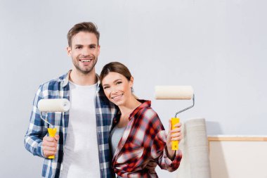 front view of happy young couple with paint rollers looking at camera while hugging at home clipart