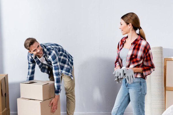 smiling young woman with gloves looking at man holding cardboard boxes at home