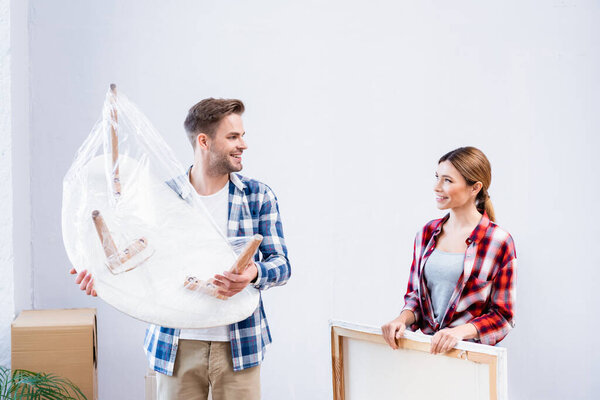 smiling young man with coffee table covered with polyethylene looking at woman holding picture indoors