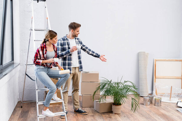 young man pointing with hand while holding coffee cup near woman sitting on ladder during repair at home