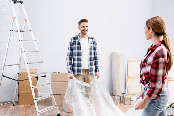 smiling young man looking at woman while covering room with polyethylene during repair at home